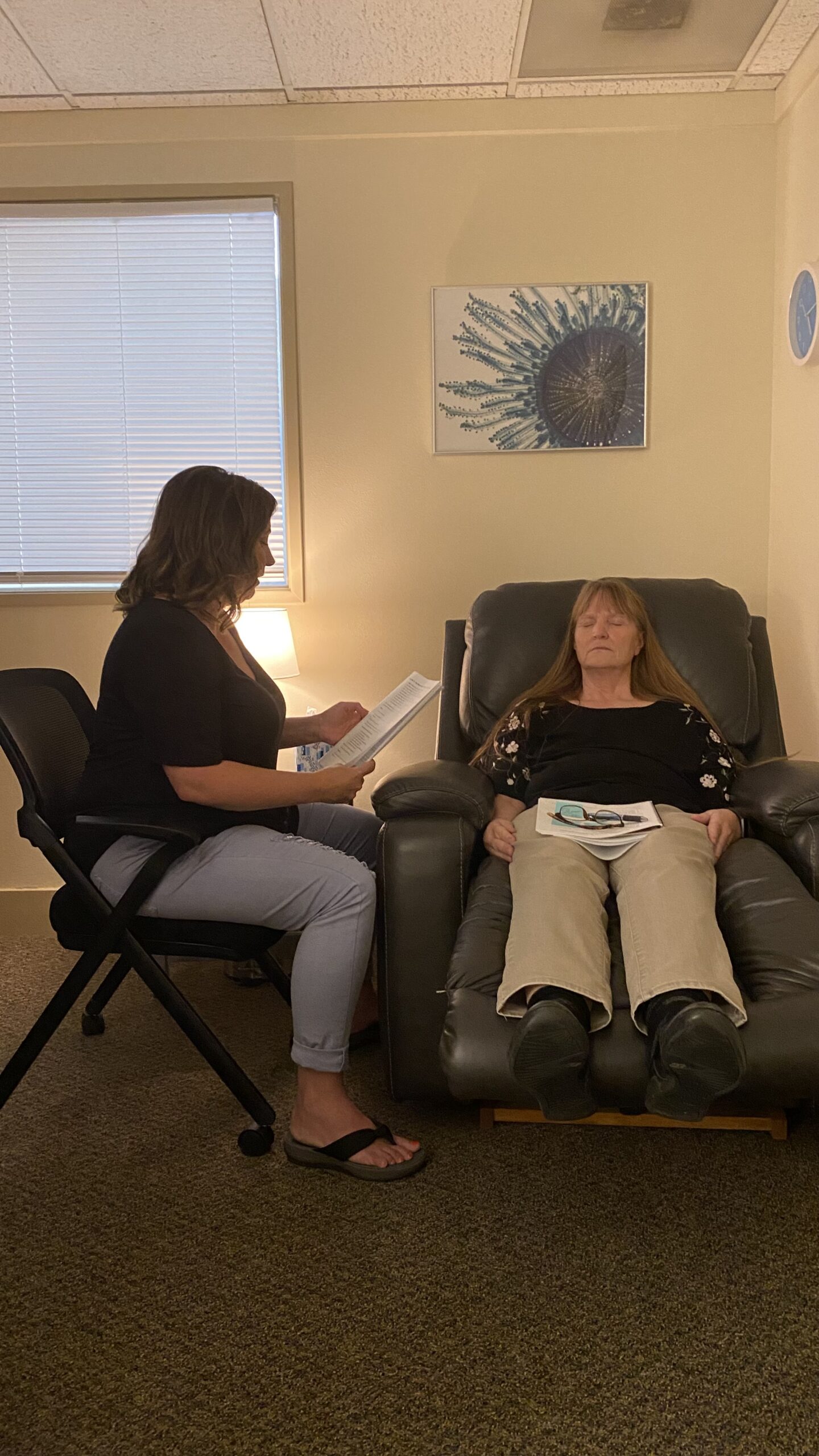 In a small office, a woman reclines with her eyes closed in a leather armchair with papers and glasses on her lap while another woman sits beside her reading from a document.