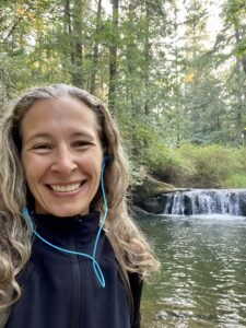 Smiling woman with long wavy hair wearing blue earbuds and a dark jacket stands beside a small cascading waterfall and forested stream.