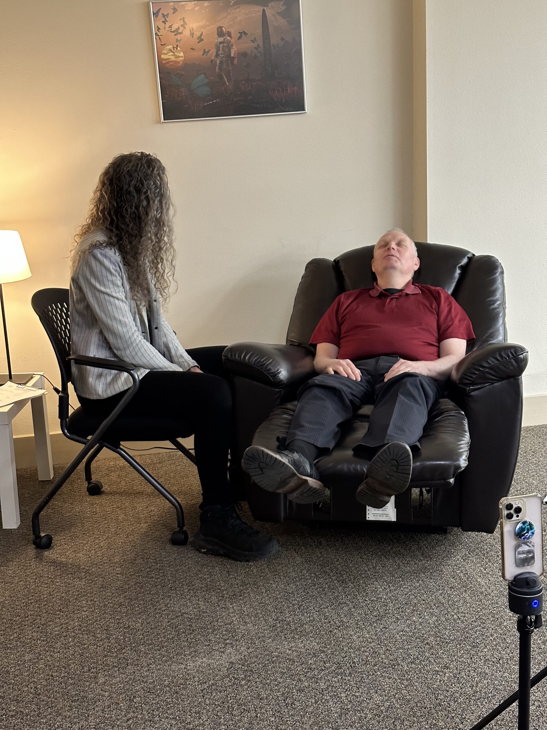 A man in a maroon polo reclines with his eyes closed in a black leather recliner during a hypnosis session while a woman with long curly hair sits nearby in an office chair; a small table and lamp sit to the left, framed artwork hangs on the wall behind them, and a smartphone on a tripod records at the right.