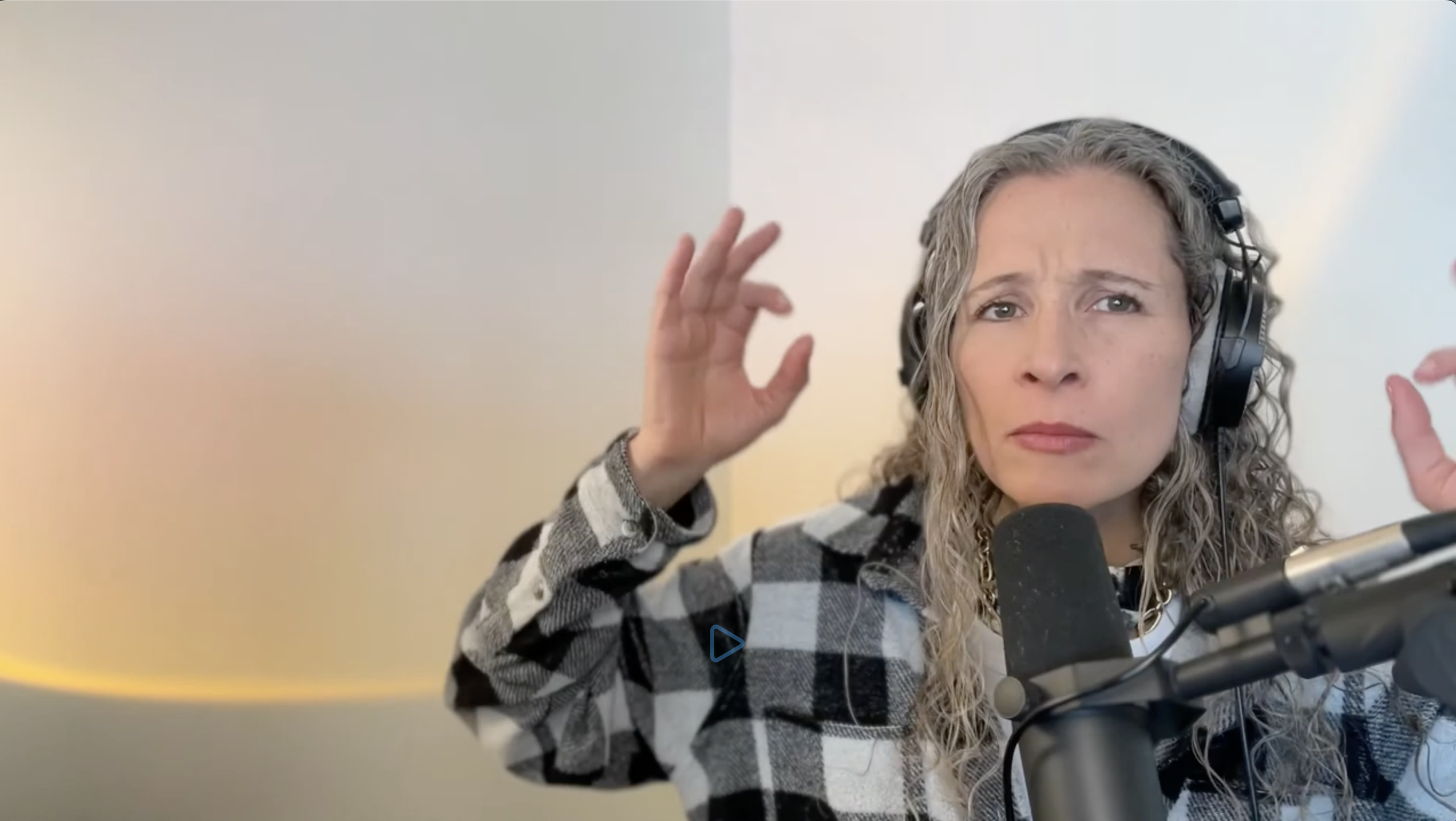 Erika Flint wearing headphones and a black-and-white plaid shirt, speaking into a microphone and gesturing with both hands while listening intently against a softly lit neutral background.