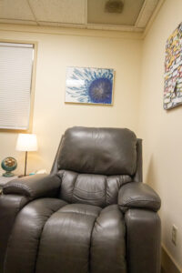 Dark brown leather recliner centered in a small therapy office, with a lit table lamp and retro fan on a side table, a framed blue abstract print on the wall, and a board of colorful notes.